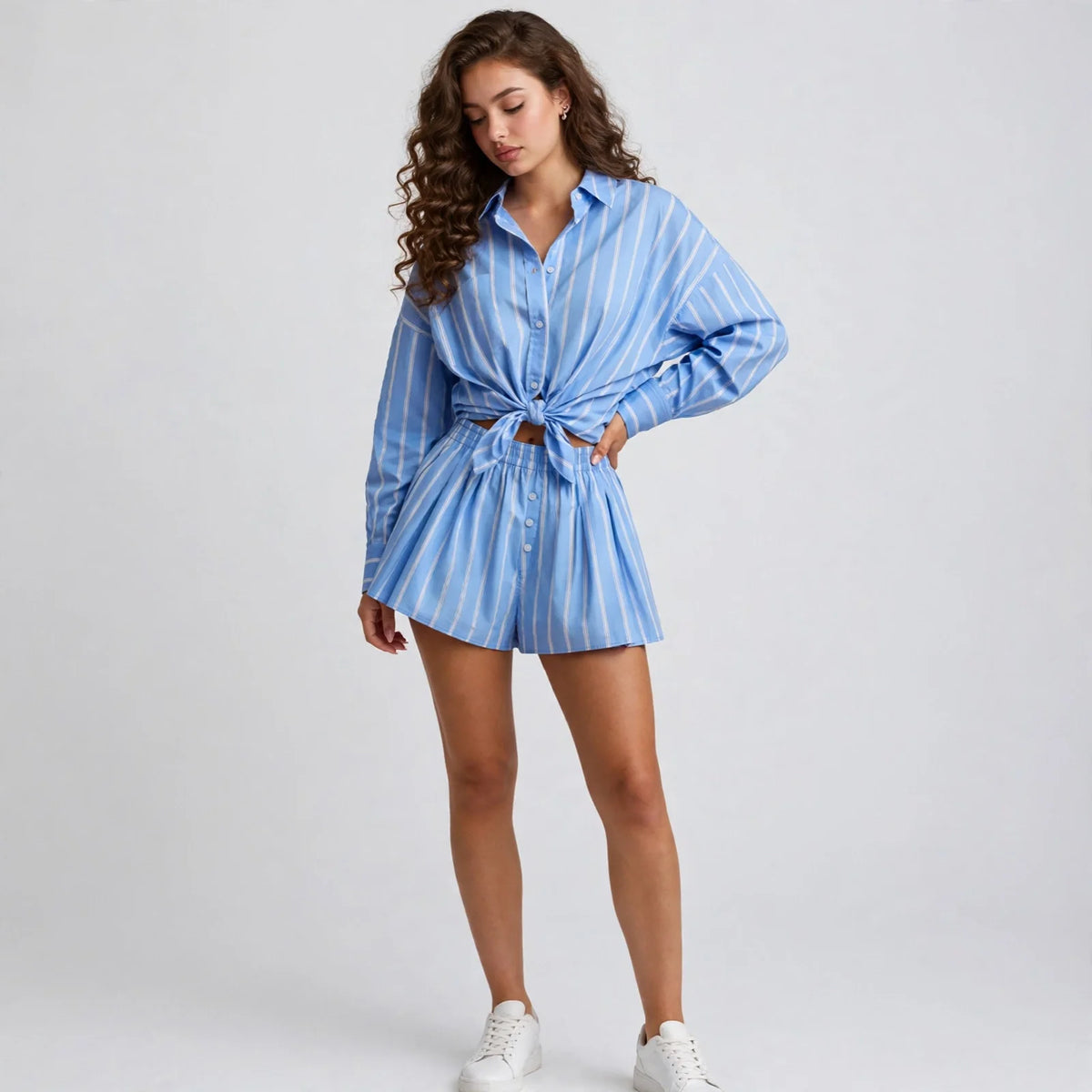 Woman in blue and white striped button-up shirt and shorts set, white sneakers, hand on hip, front view, light backdrop.