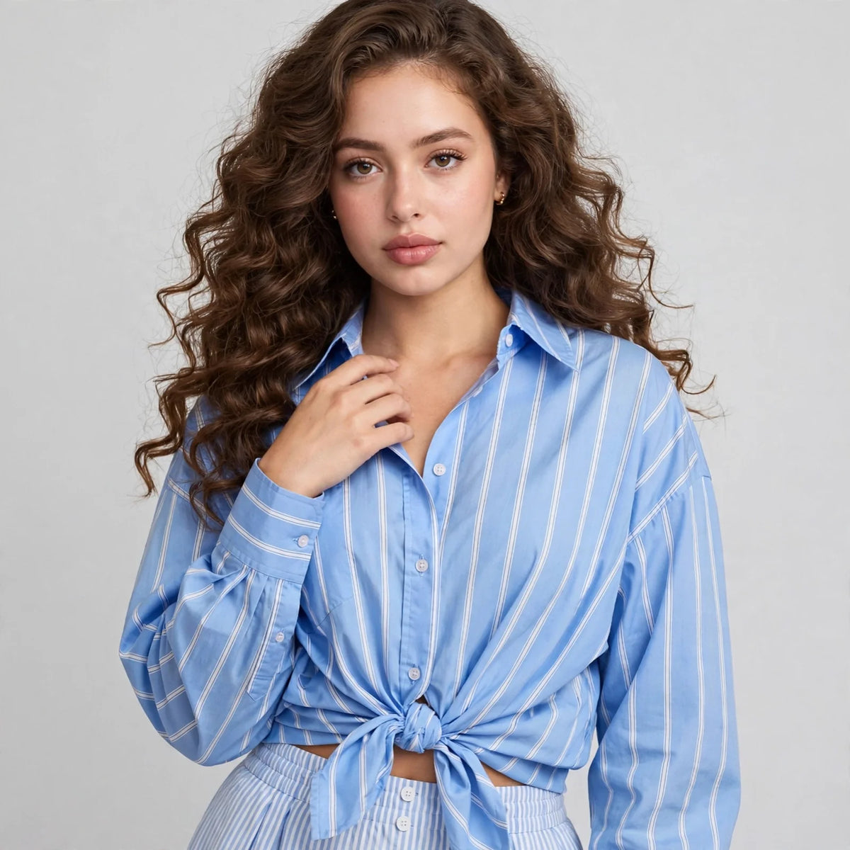 Close-up of woman in blue and white striped button-up shirt with tied front knot, collar and buttons, light studio backdrop.