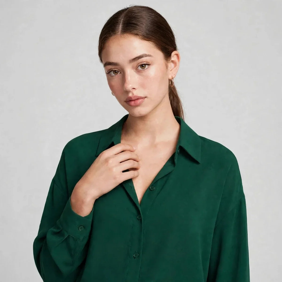 Close-up of a woman wearing a dark green button-up shirt with collar, hand at neckline, against a gray studio background.