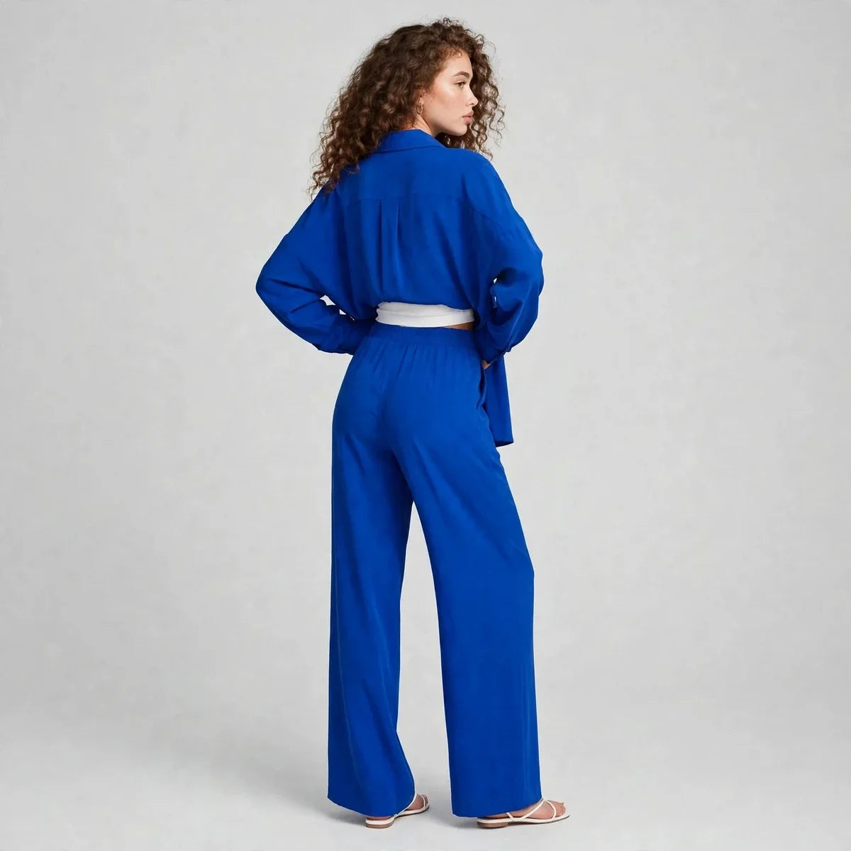 Back view of a woman wearing a royal blue matching set with long-sleeve collared shirt, wide-leg pants and white belt.