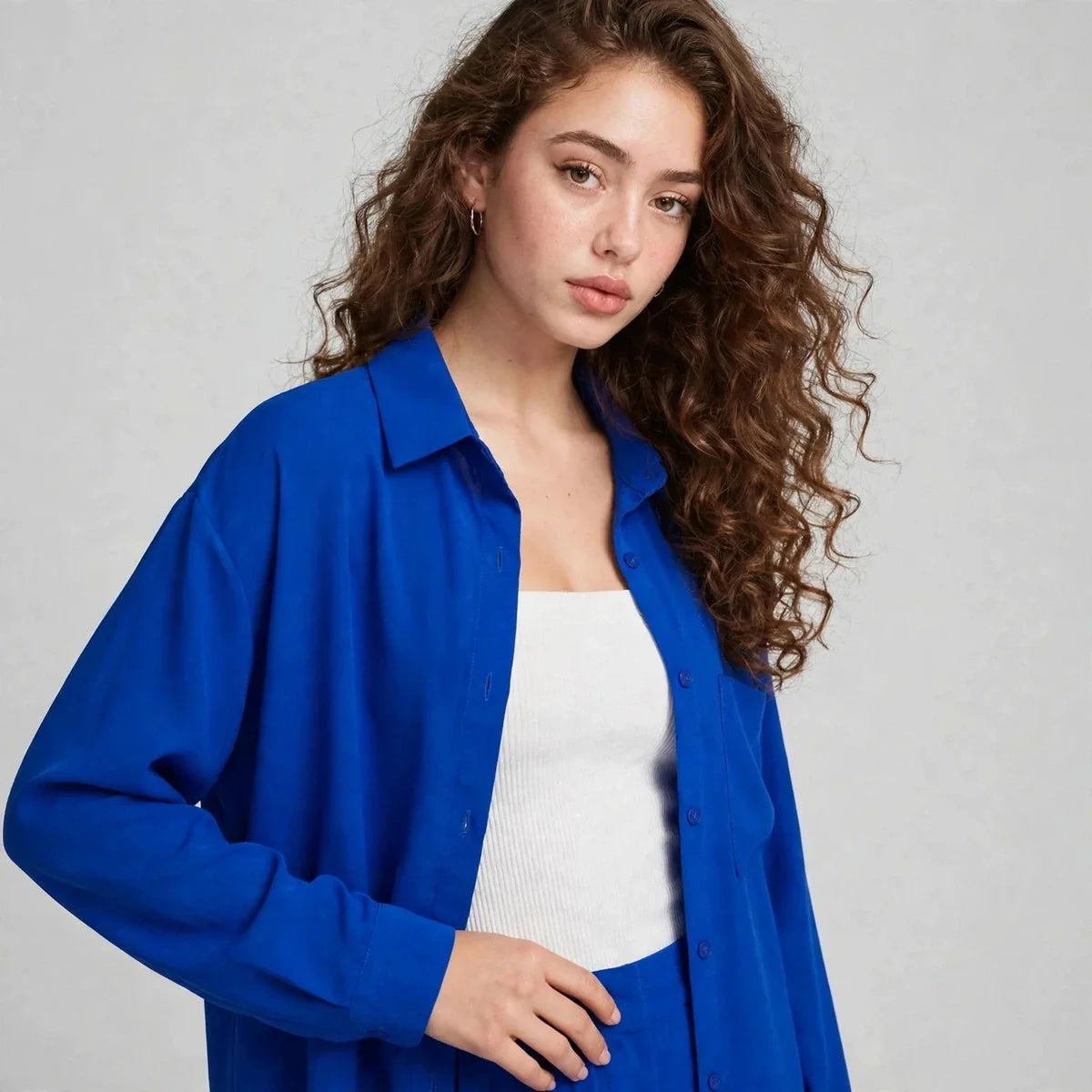 Close-up of a woman wearing a royal blue button-up shirt over a white ribbed tank top, with long curly brown hair.
