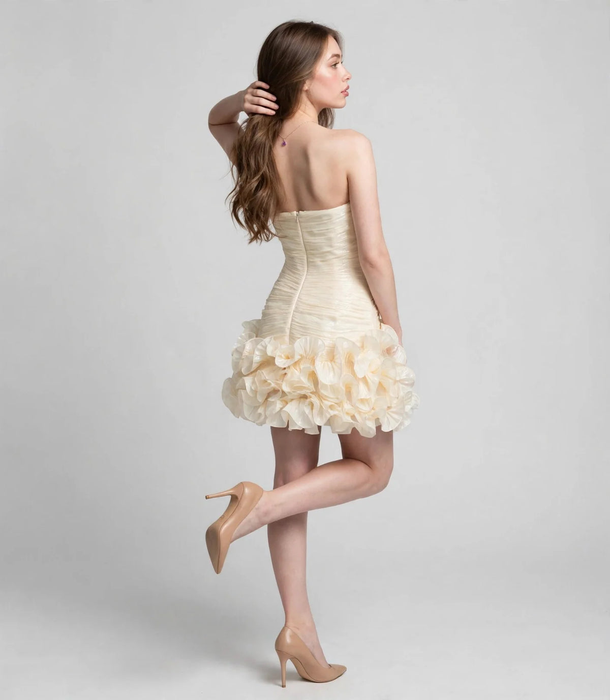 Back view of a woman wearing a cream strapless mini dress with a ruched bodice and tiered ruffle skirt, nude high heels.