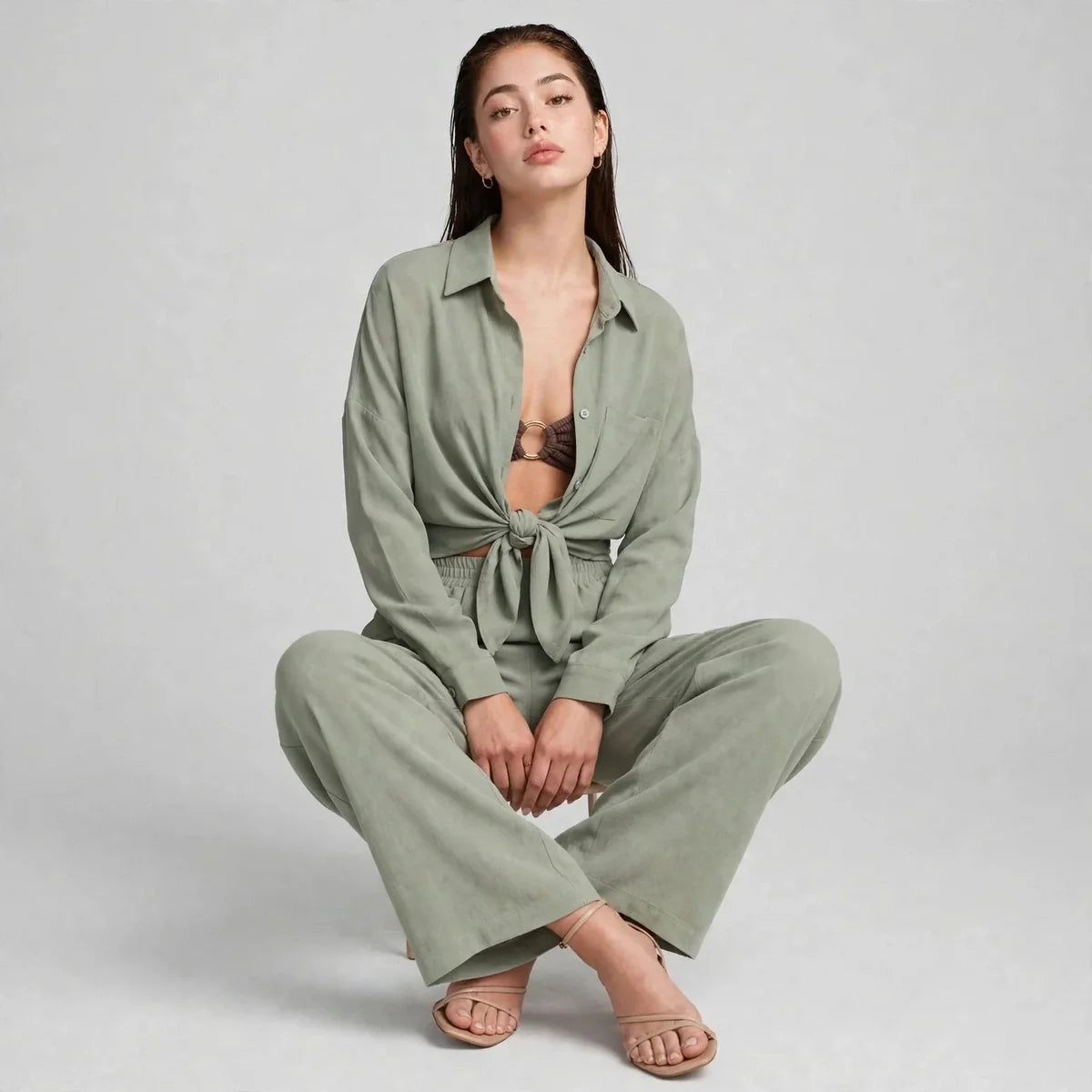 Woman crouching in a sage green matching set with button-front shirt tied at the waist, wide-leg pants and nude sandals.