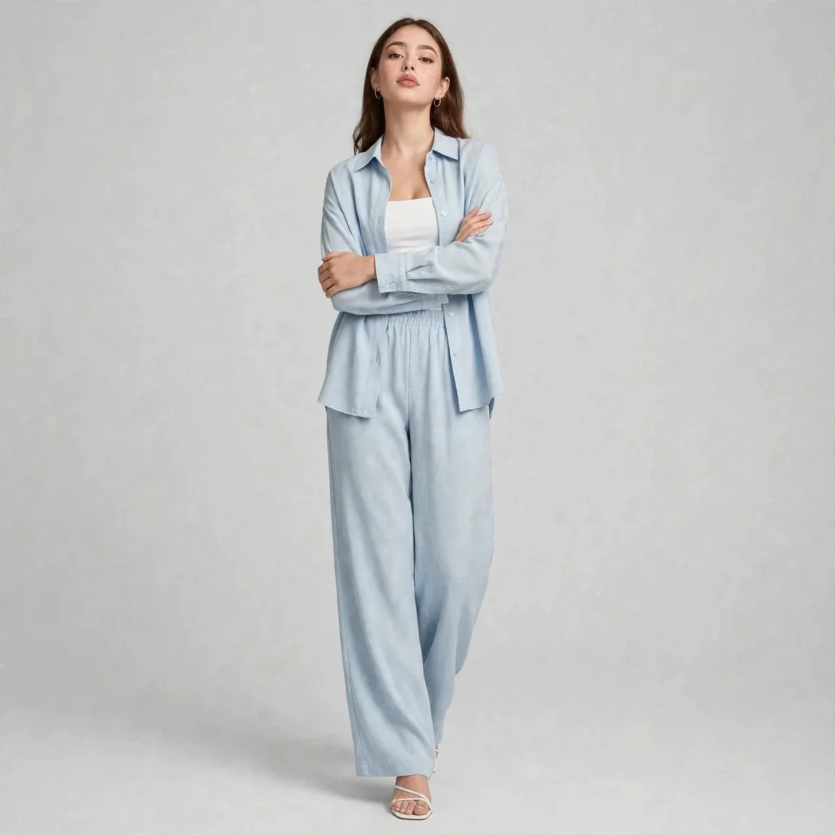 Woman with arms crossed modeling a light blue two-piece set with long-sleeve button-up shirt over a white tank top.
