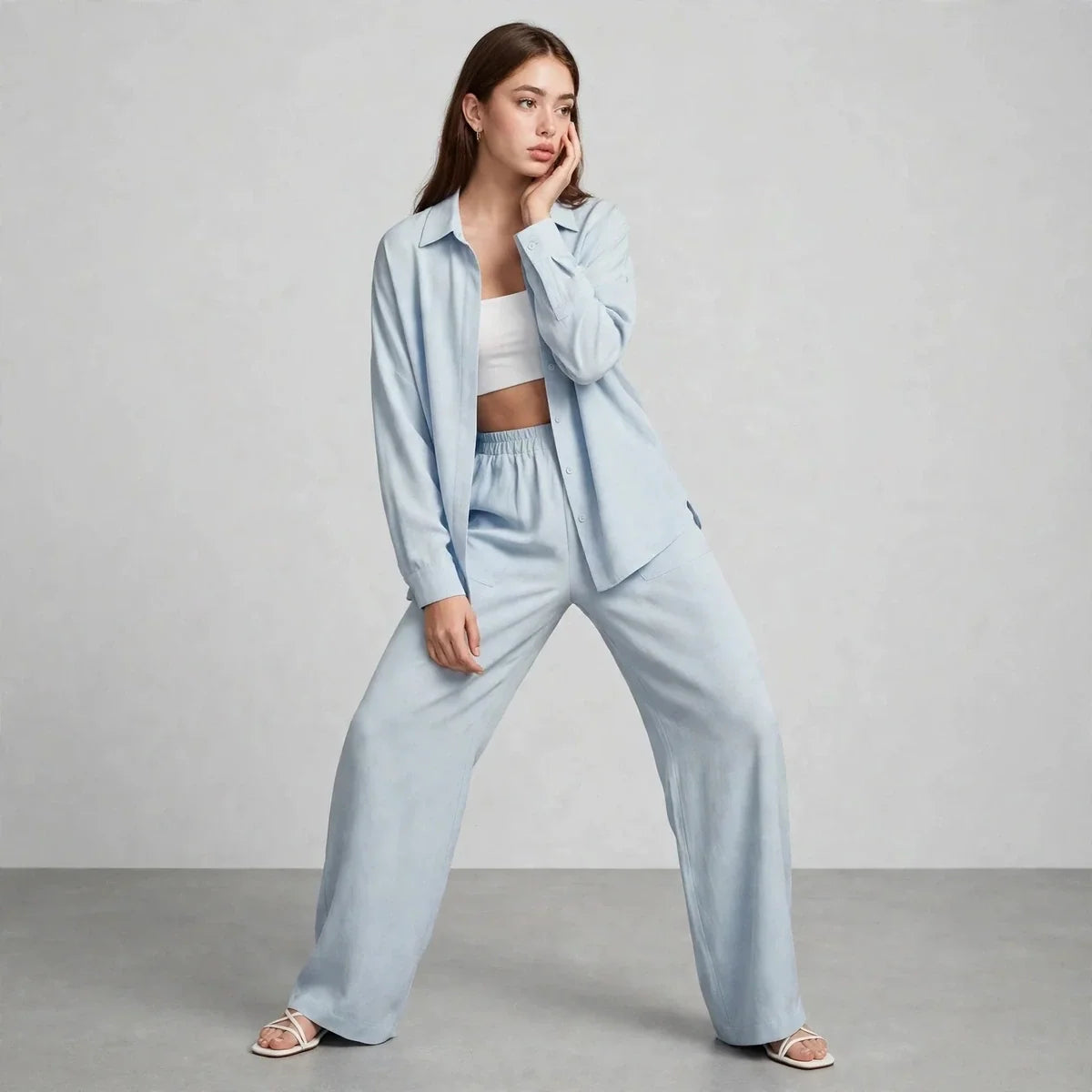 Woman modeling a light blue two-piece set with long-sleeve button-up shirt worn open over a white crop top, wide-leg pants.
