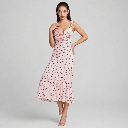 Woman in a pink heart-print midi dress with draped cowl neckline and ruffled hem, spaghetti straps, gray background.