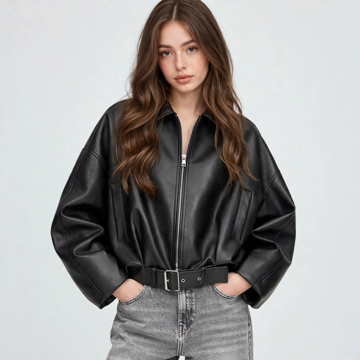 Front view of model with hands in pockets wearing a black cropped faux leather zip-up jacket with belted hem and grey jeans