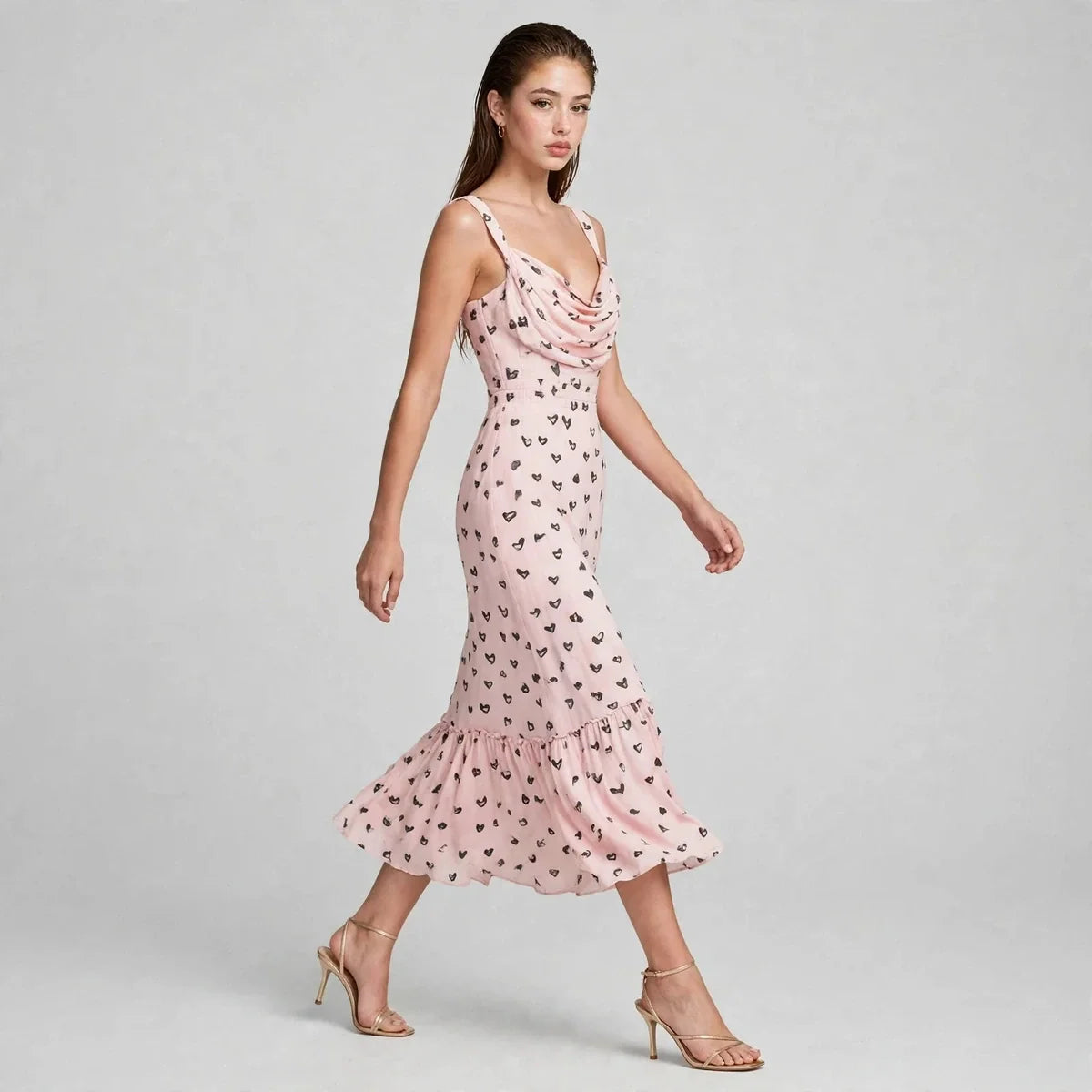 Woman walking in pink heart-print midi dress with draped neckline and ruffled hem, side view, heels, gray background.