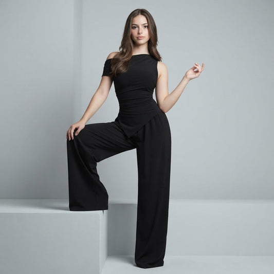 Woman posing with leg on a platform in a black one-shoulder top and matching wide-leg pants, gray studio backdrop.