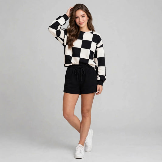 Model in black and white checkerboard knit sweatshirt with black drawstring shorts, white sneakers, full-length studio shot.