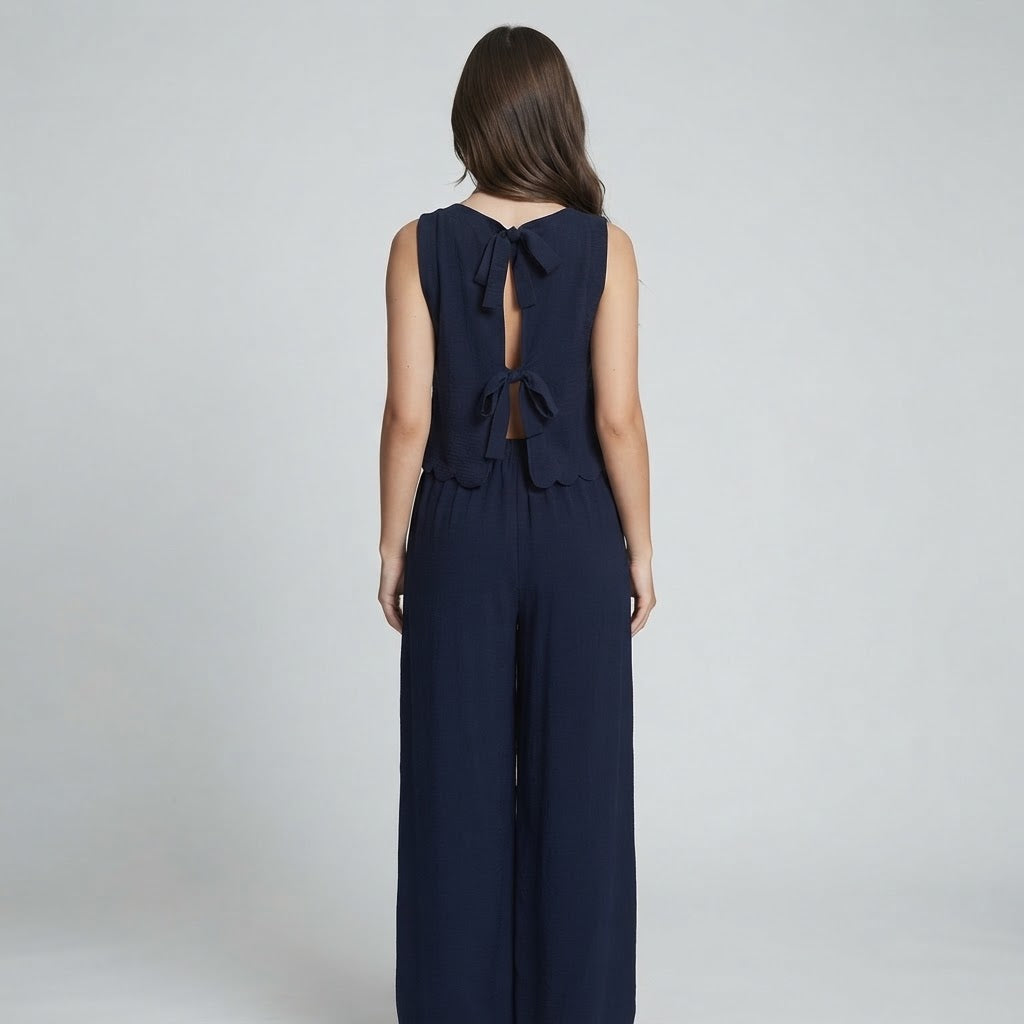 Back view of a woman wearing a navy sleeveless top with tie-back detail and matching wide-leg pants, gray studio background.