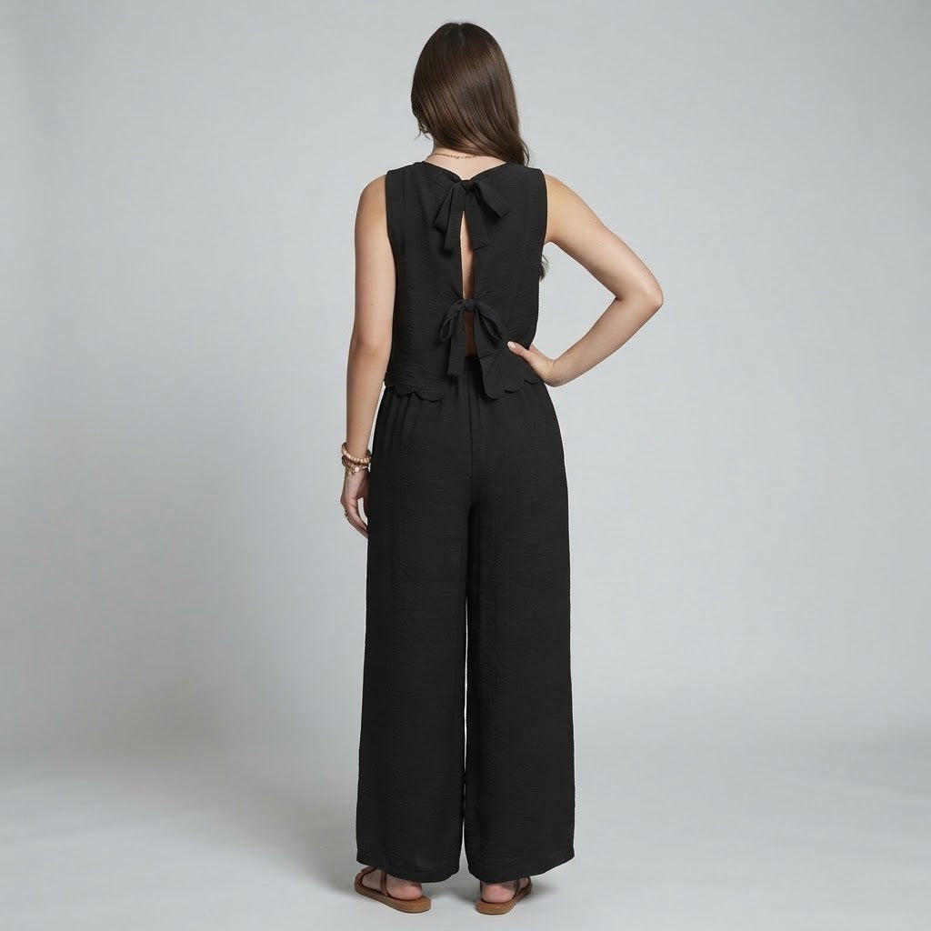 Back view of a woman wearing a black sleeveless top with bow tie-back detail and matching wide-leg pants, gray background.