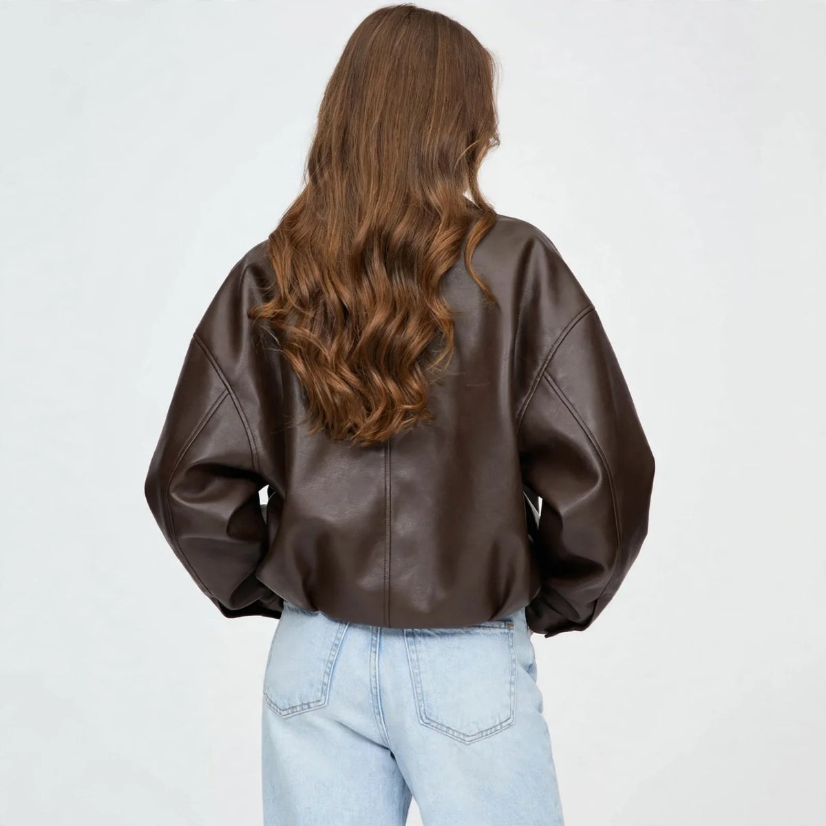 Back view of model wearing a brown cropped faux leather zip-up jacket with belted hem and light-wash jeans, grey background
