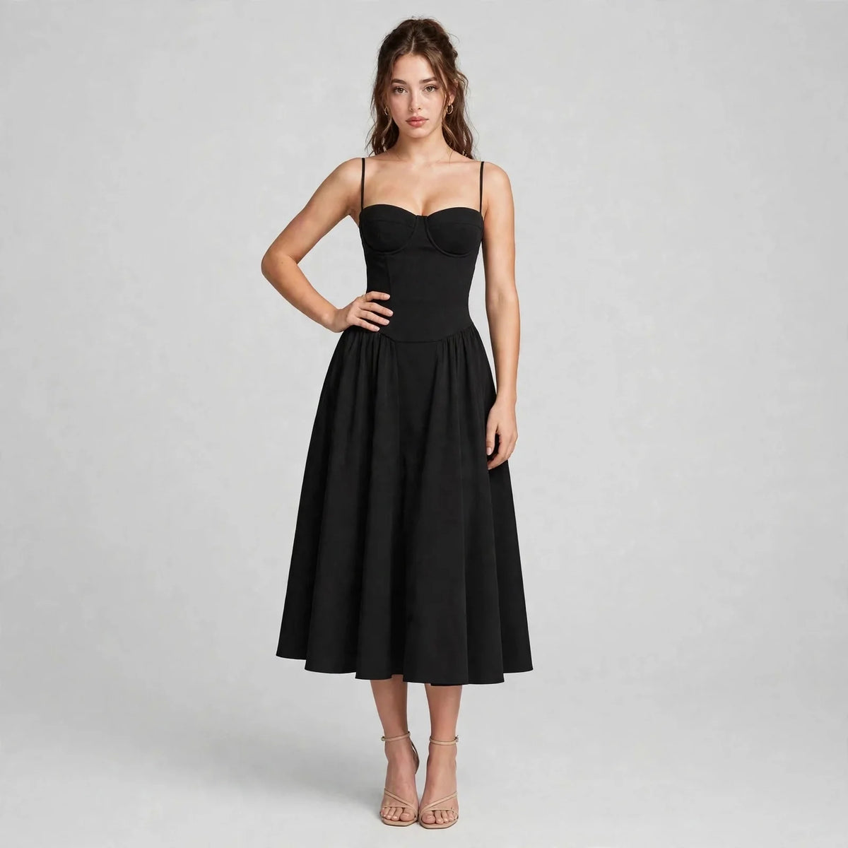 Front view of model with hand on hip wearing a black sweetheart neck ruched midi dress with spaghetti straps, grey background
