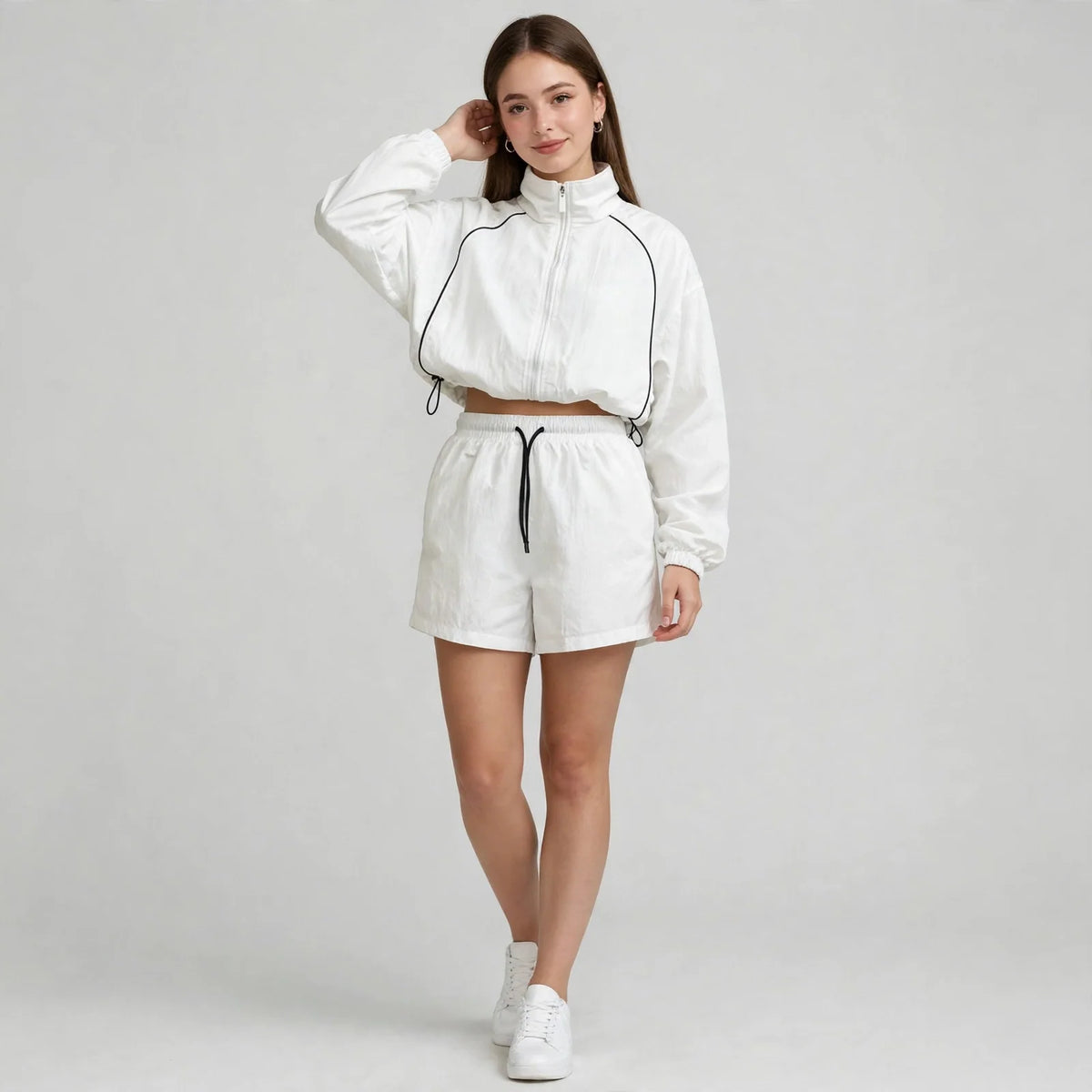 Woman in a white cropped zip-up jacket and matching shorts with black piping and drawstrings, front view, gray background.