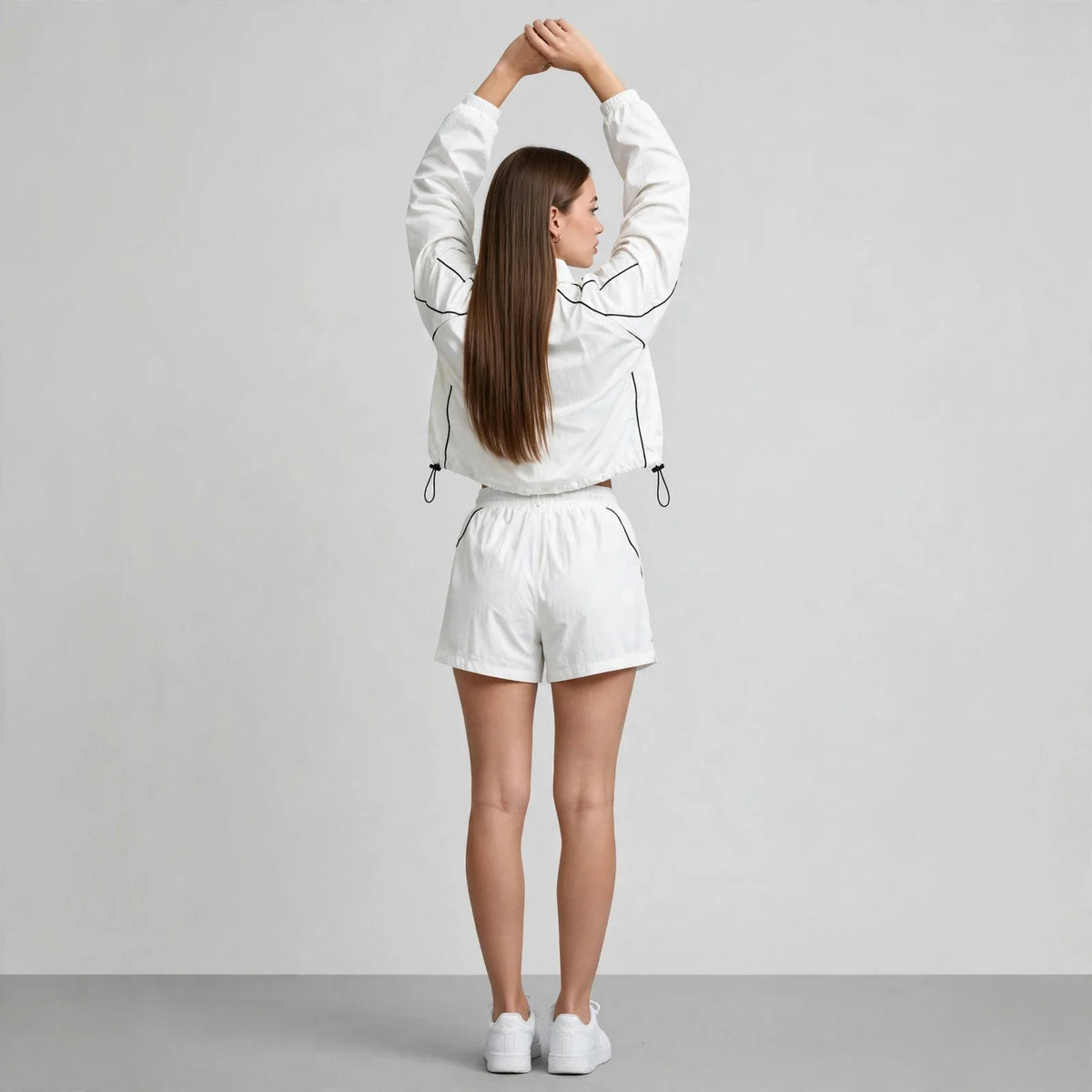 Back view of a woman in a white cropped zip-up jacket and matching shorts, arms raised overhead, gray studio background.