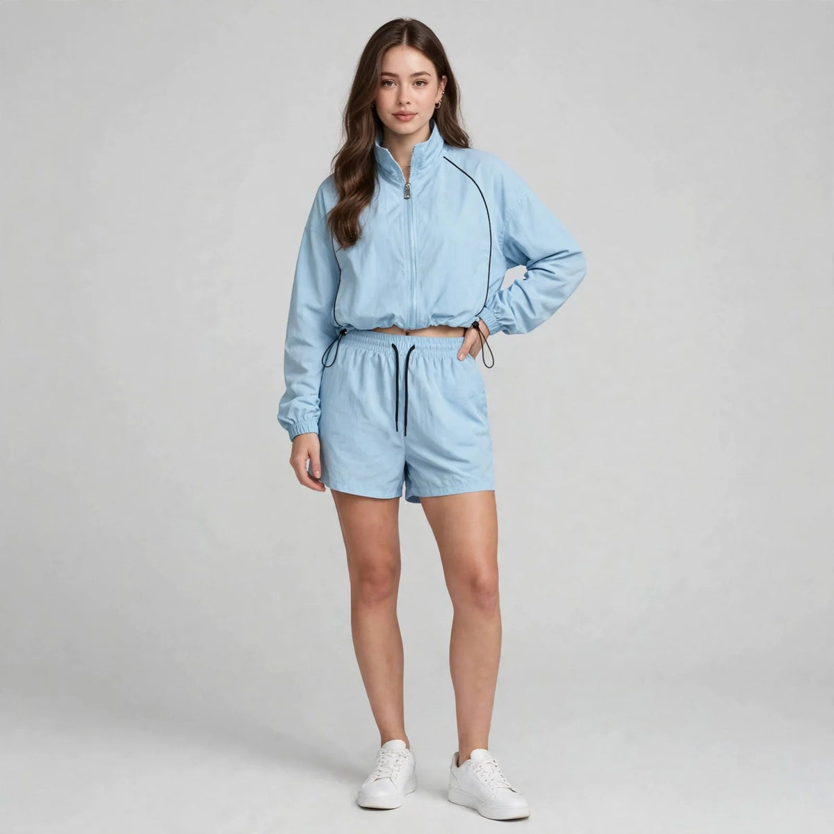 Woman in a light blue zip-up jacket and matching shorts with white sneakers, front view, light gray studio background.
