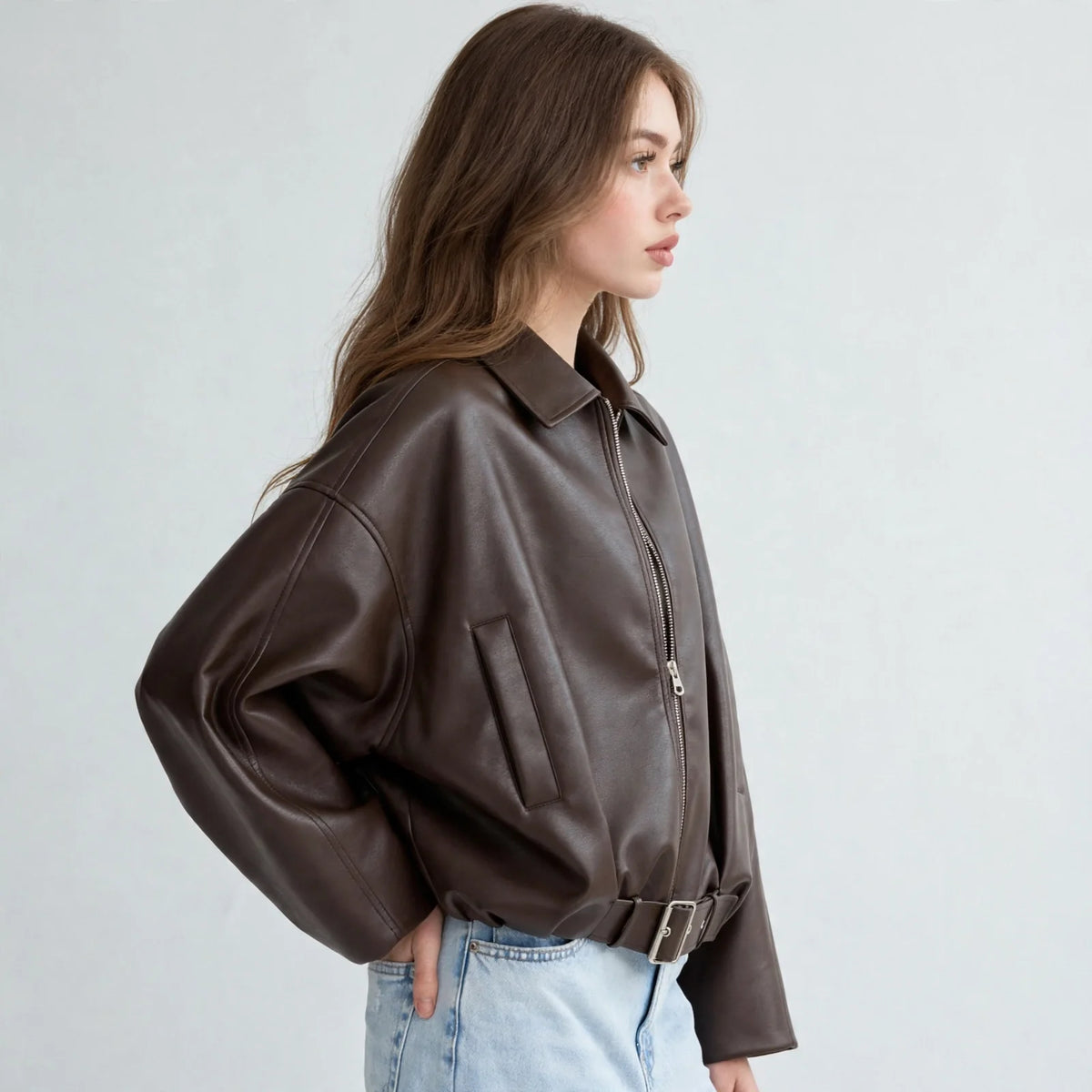 Side view of model wearing a brown cropped faux leather zip-up jacket with belted hem and light-wash jeans, grey background