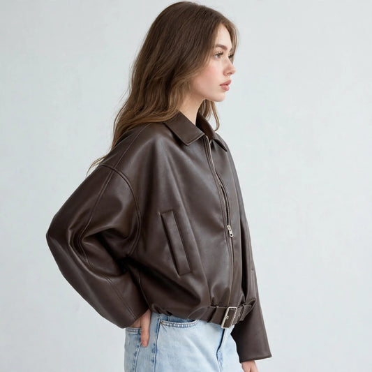 Side view of model wearing a brown cropped faux leather zip-up jacket with belted hem and light-wash jeans, grey background