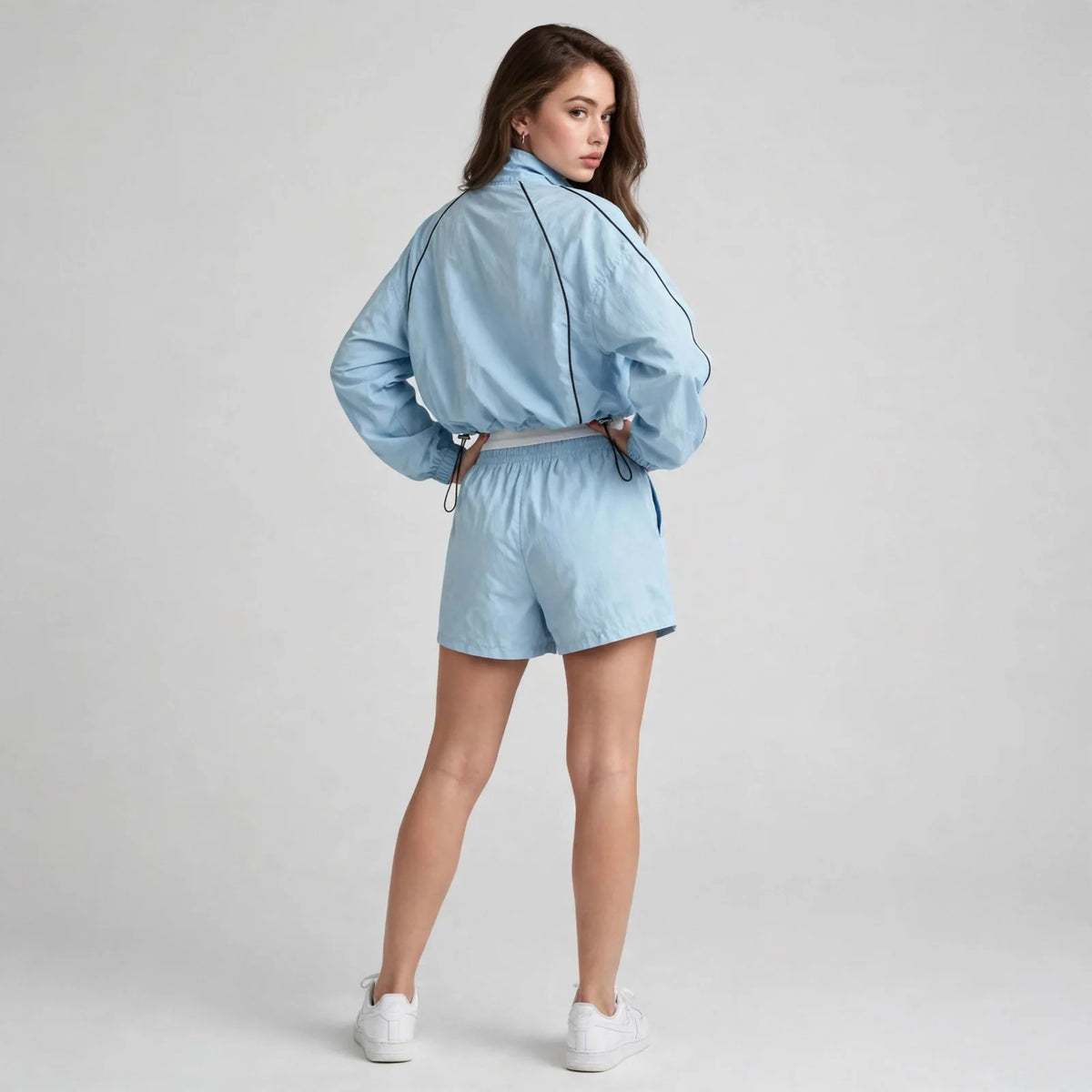 Back view of a woman in a light blue cropped zip-up jacket and matching shorts with white sneakers, gray studio background.