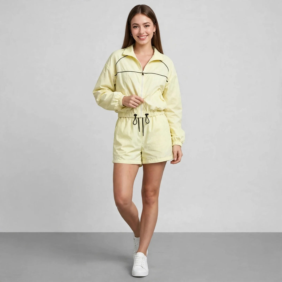 Woman in a pale yellow cropped zip-up jacket and matching shorts with dark piping, walking pose, gray studio background.