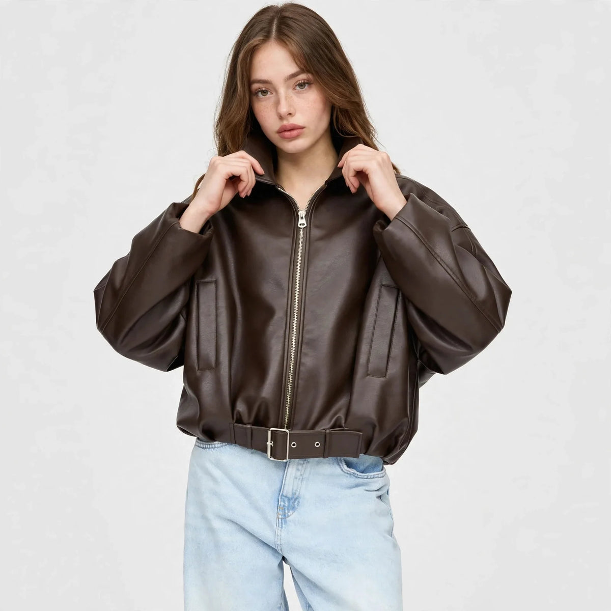 Front view of model with hands at collar wearing a brown cropped faux leather zip-up jacket with belted hem and light jeans