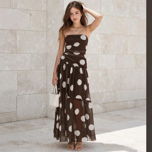 Woman wearing a brown strapless maxi dress with large white polka dots, holding a white bag, beige stone wall background.