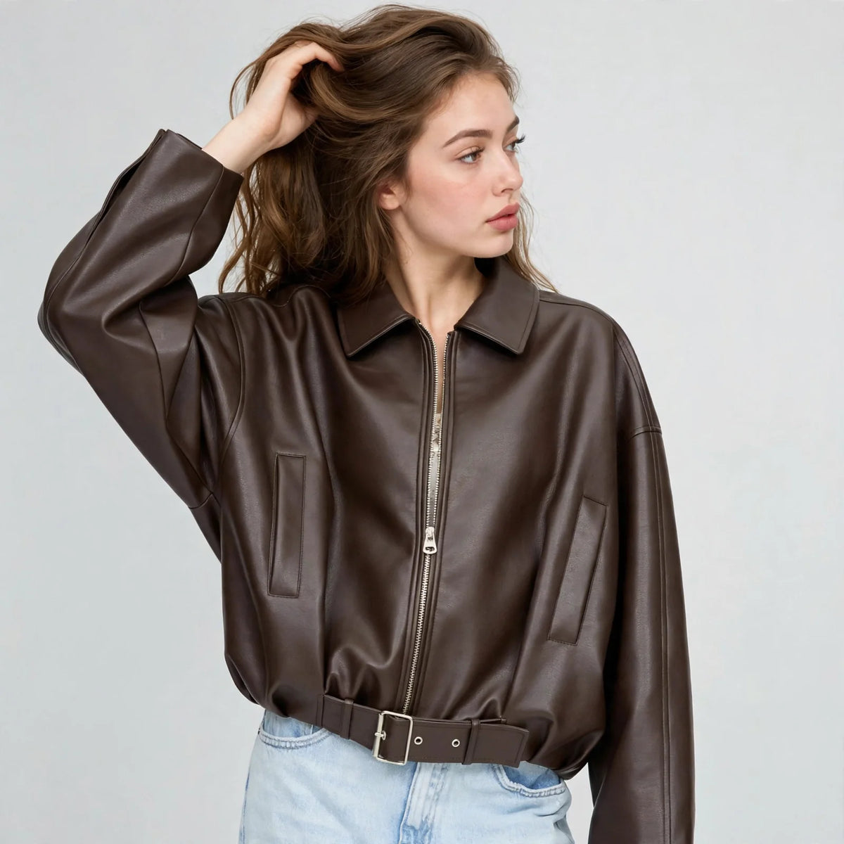 Front view of model with hand in hair wearing a brown cropped faux leather zip-up jacket with belted hem, grey background