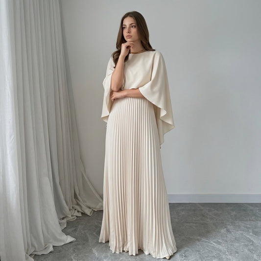 Front view of a woman in an ivory cape-style maxi dress with draped cape sleeves and pleated skirt, white curtains behind.