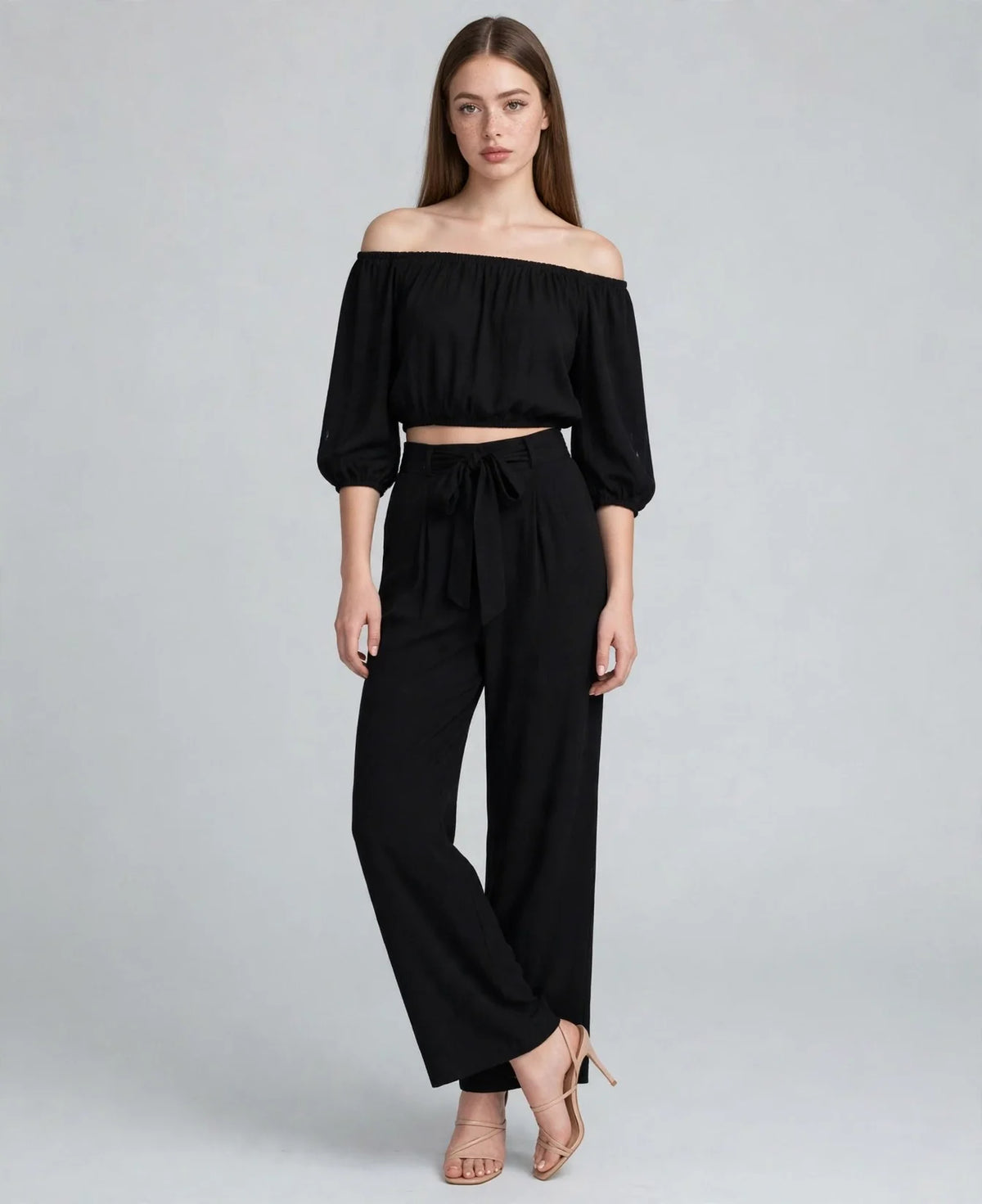 Standing model in a black off-shoulder two-piece set, 3/4 puff sleeves, cropped top, tie-waist wide-leg pants, grey backdrop.