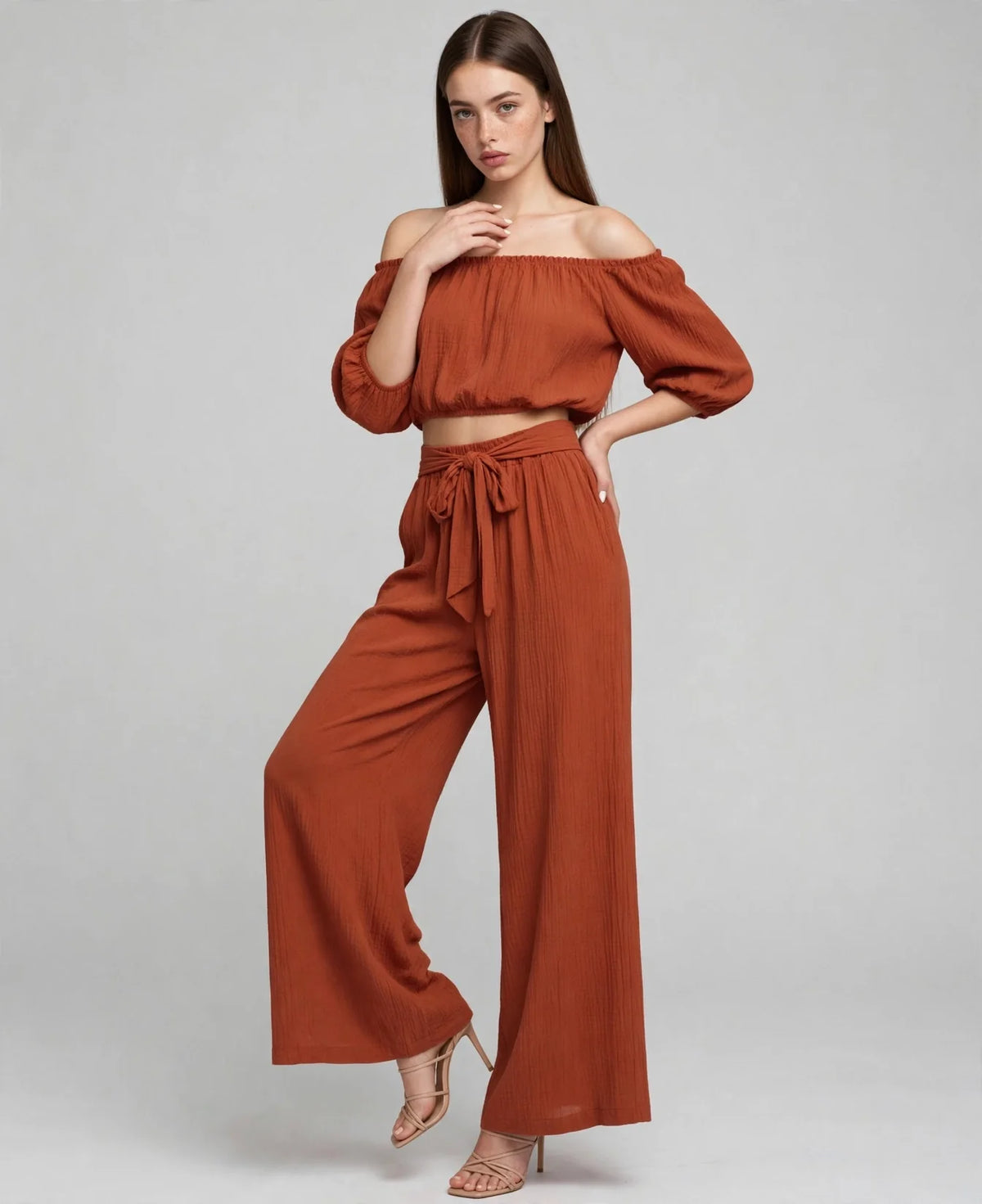 Model walking in a rust off-shoulder two-piece set, puff-sleeve crop top and wide-leg tie-waist pants, grey studio backdrop.
