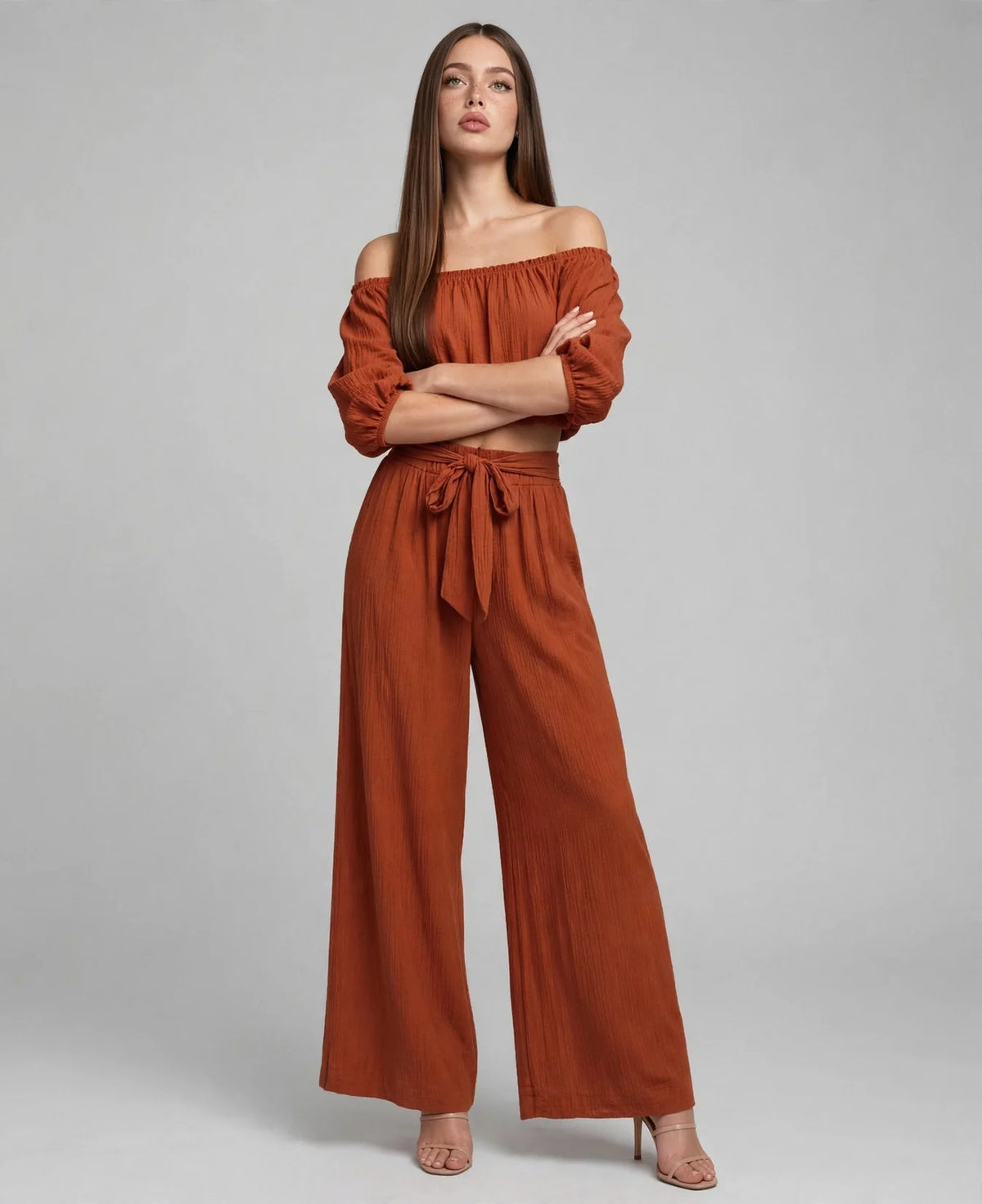 Model with crossed arms in a rust off-shoulder cropped top and matching wide-leg tie-waist pants, grey studio background.