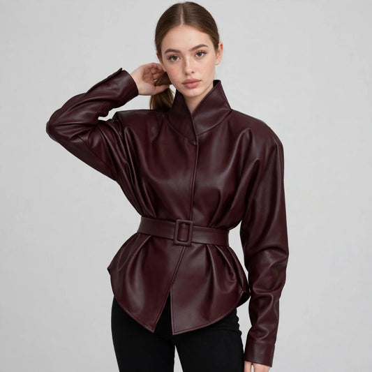 Woman in a burgundy faux leather jacket with stand collar and belted waist, black pants, gray studio background.