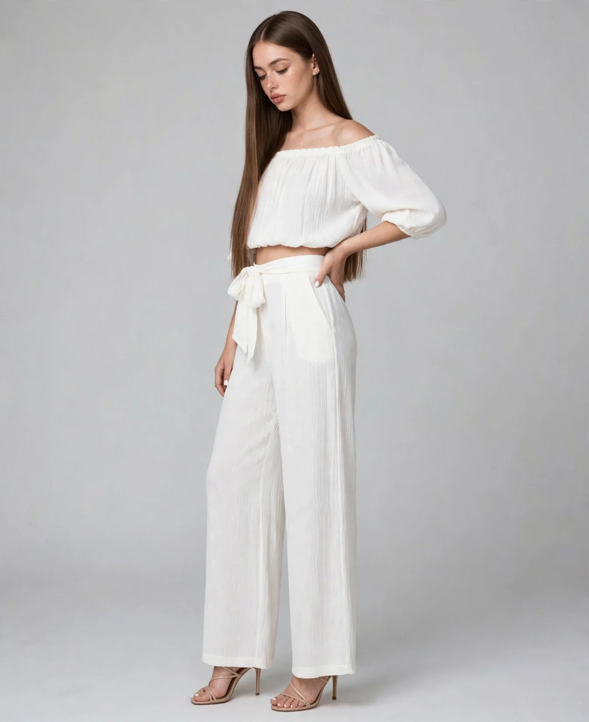 Model in a white off-shoulder crop top and matching wide-leg tie-waist pants, standing pose on light grey studio background.