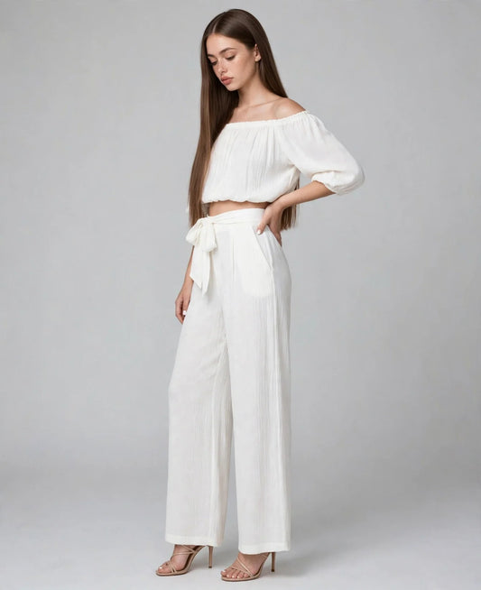 Model in a white off-shoulder crop top and matching wide-leg tie-waist pants, standing pose on light grey studio background.