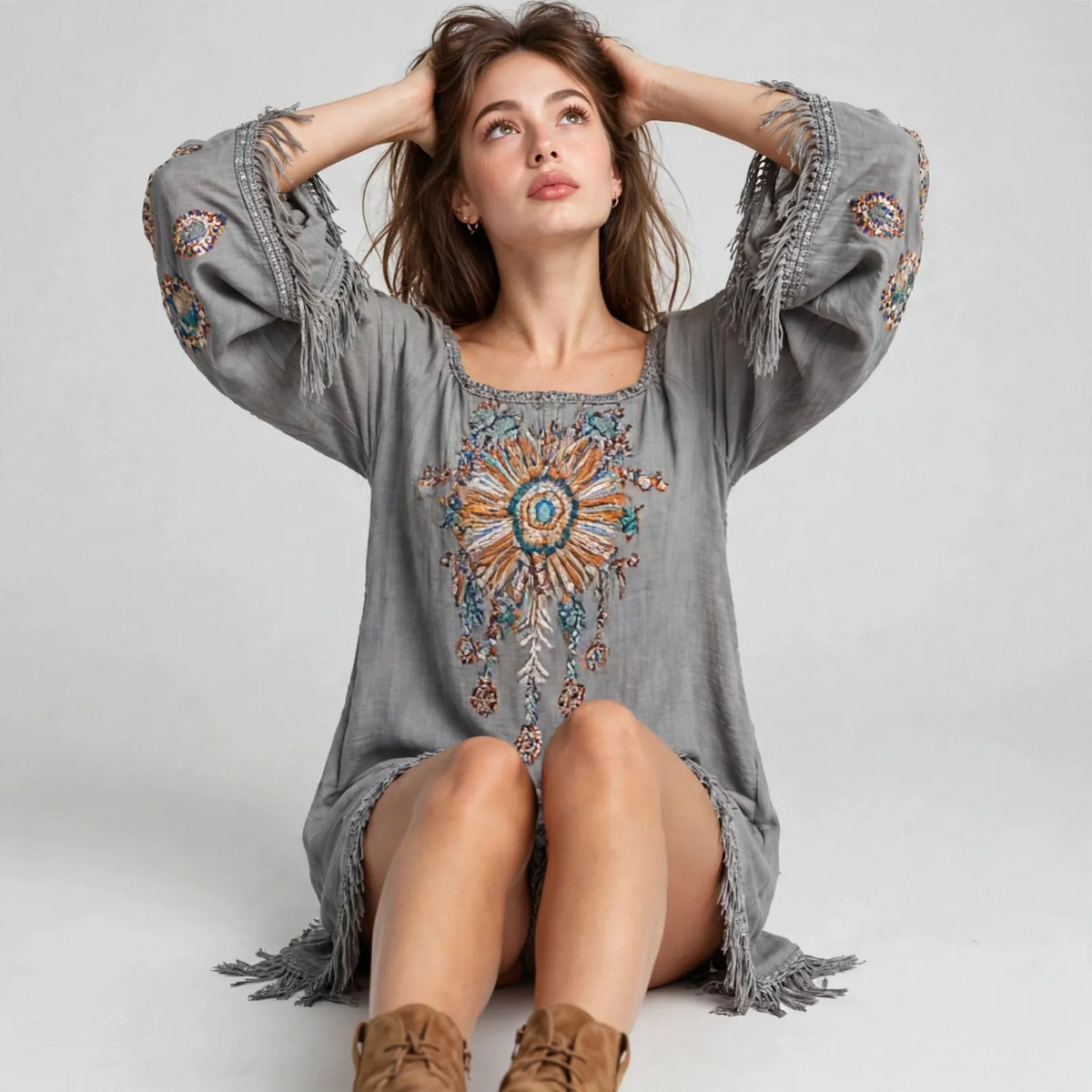 Seated model in a grey embroidered mini dress with fringed hem and bell sleeves, hands behind head, white studio background.