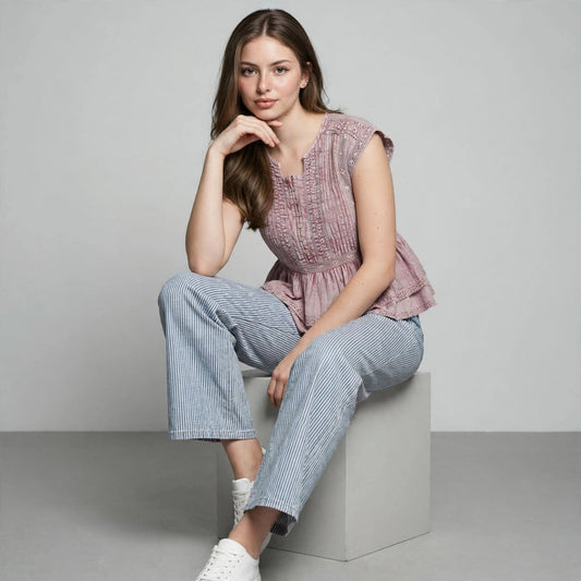 Woman sitting on a gray block wearing a dusty pink sleeveless peplum top, striped cropped pants and white sneakers.