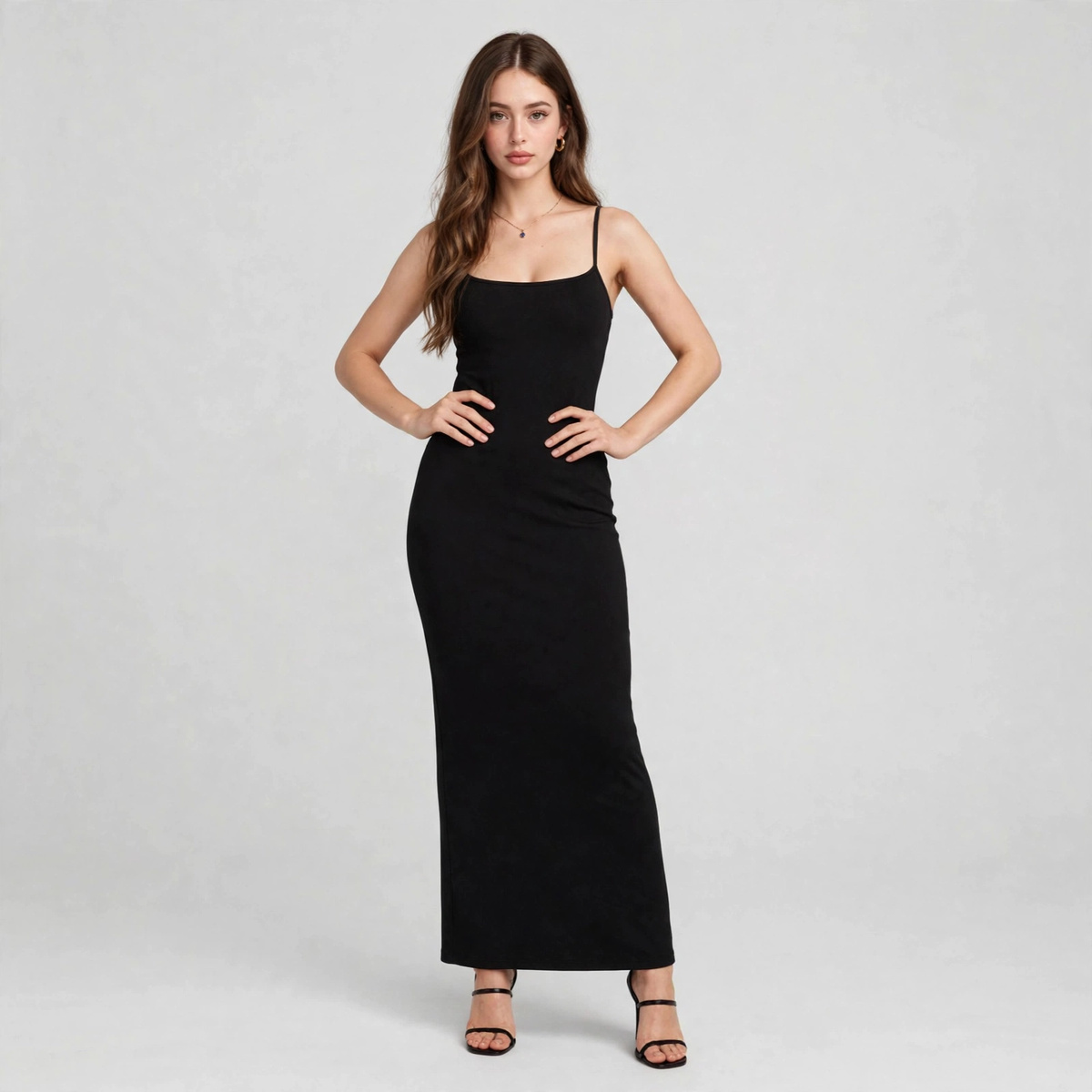 Woman standing with hands on hips in a black spaghetti-strap maxi dress with a fitted shapewear silhouette, gray background.