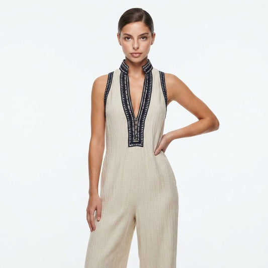 Woman in a sleeveless ivory wide-leg jumpsuit with navy embroidered V-neck trim, hand on hip, white studio background.
