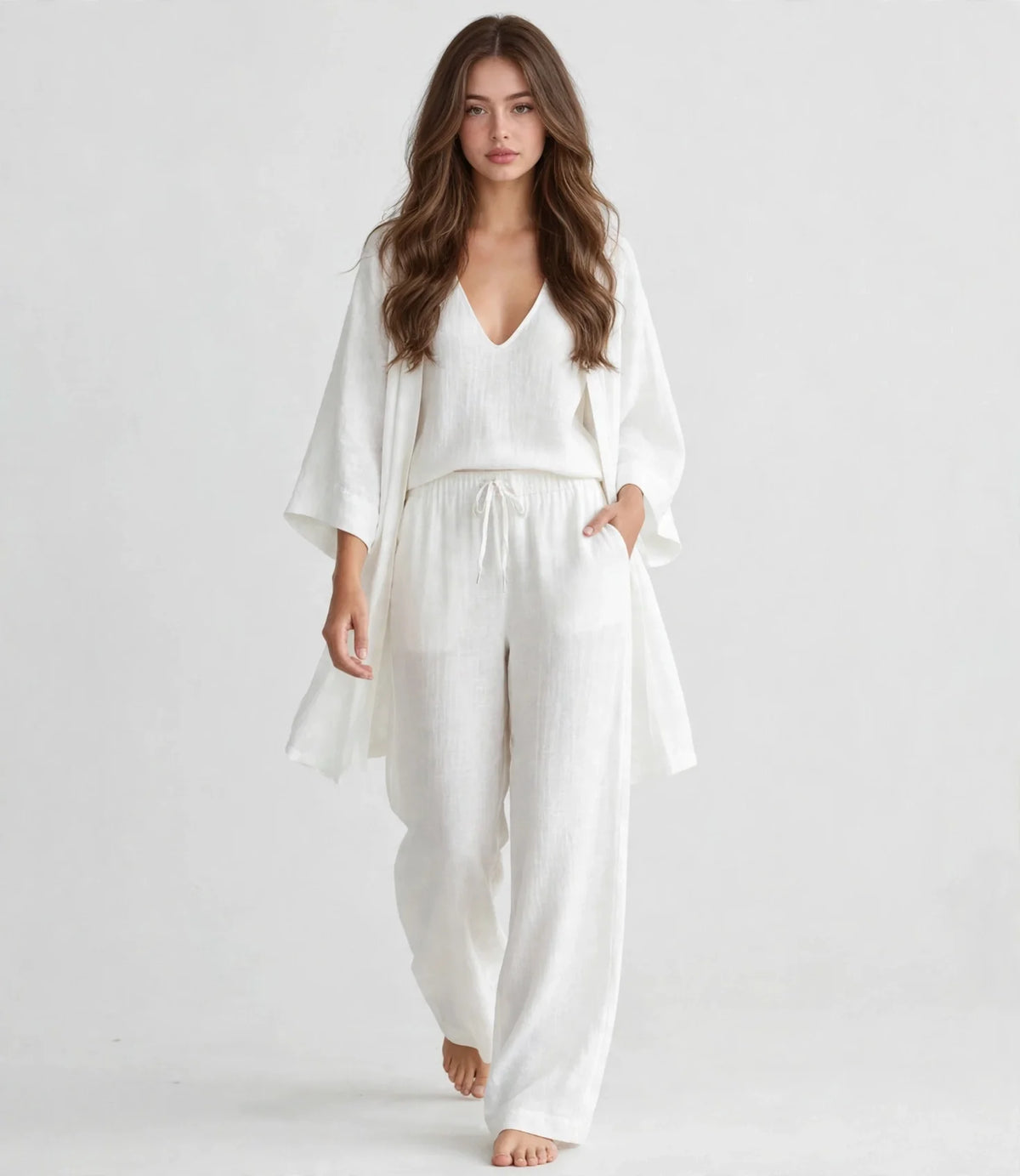 Woman in a white V-neck top, open cardigan and drawstring pants set, walking full-length pose, light gray studio background.