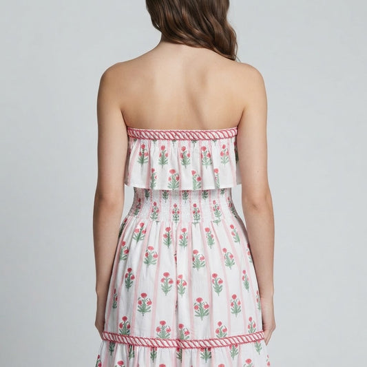Back view of a woman in a white strapless floral maxi dress with pink trim and tiered skirt, gray studio background.