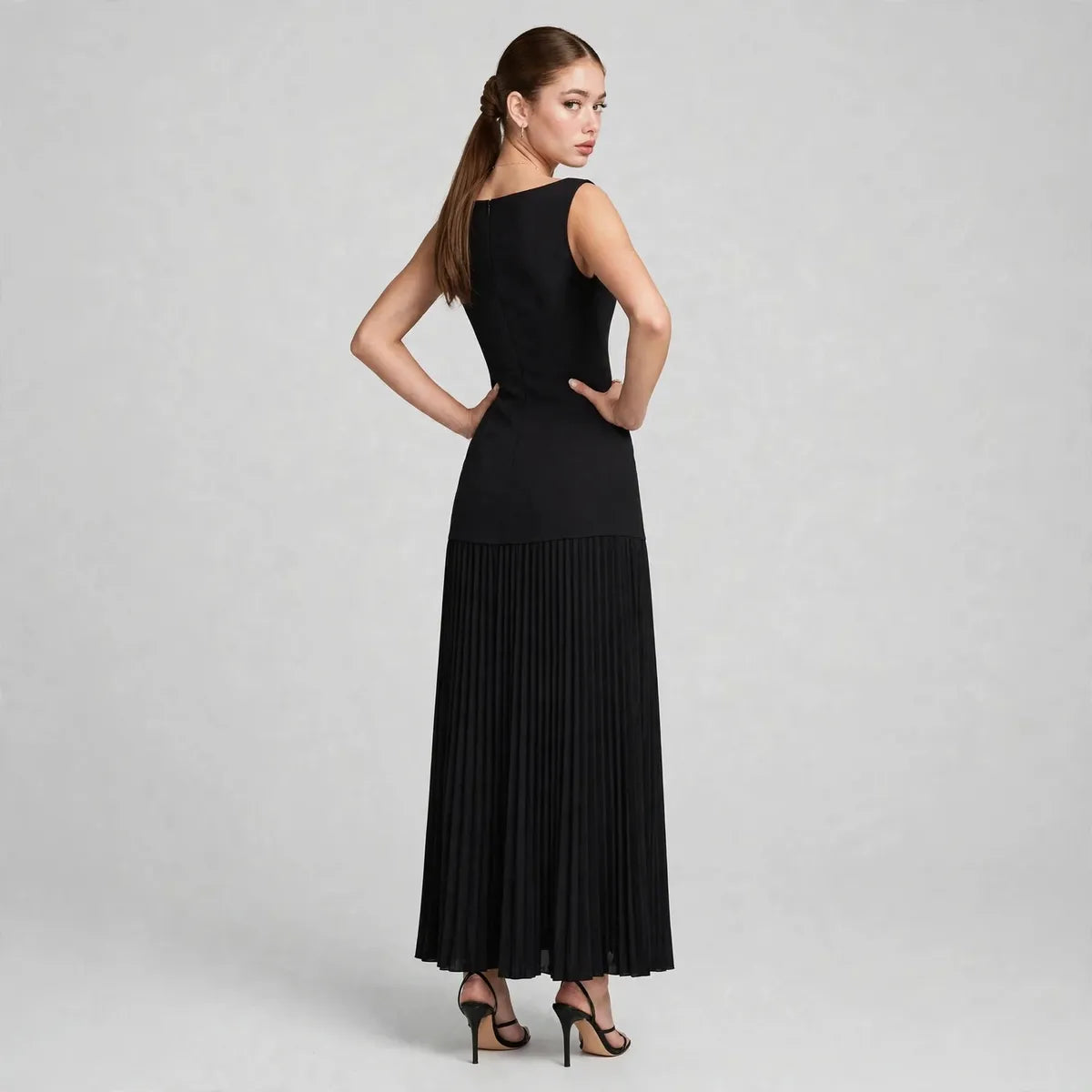 Back view of a black sleeveless pleated maxi dress with boat neckline, model standing with hands on hips in studio.