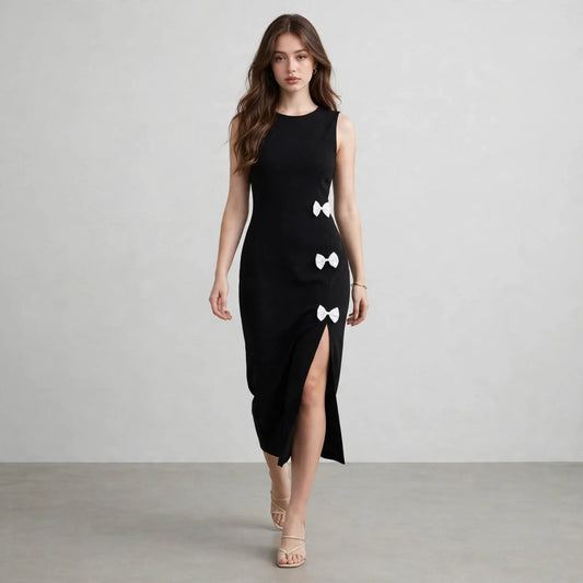 Model walking in a black sleeveless midi dress with round neck, side slit and white bow details, front view, grey background