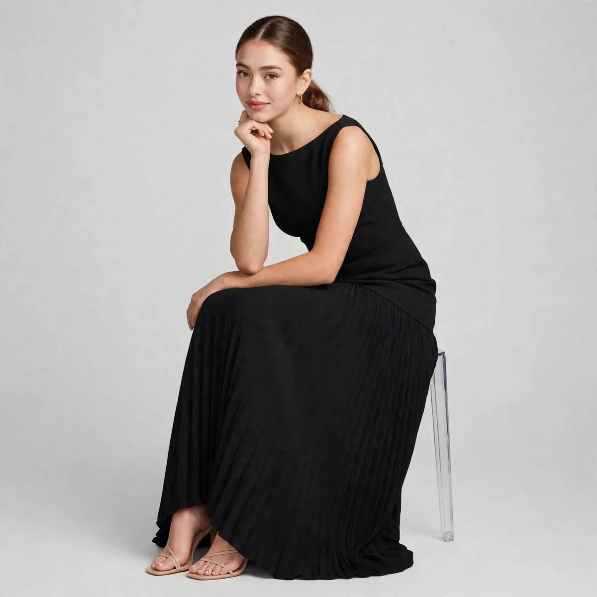 Model seated on a clear acrylic chair wearing a black sleeveless pleated maxi dress, chin resting on hand, studio.