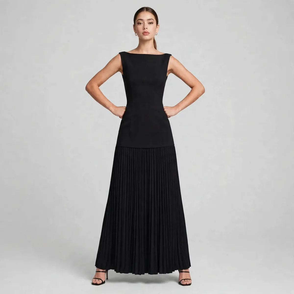 Front view of a black sleeveless maxi dress with boat neckline and pleated skirt, model posing with hands on hips.