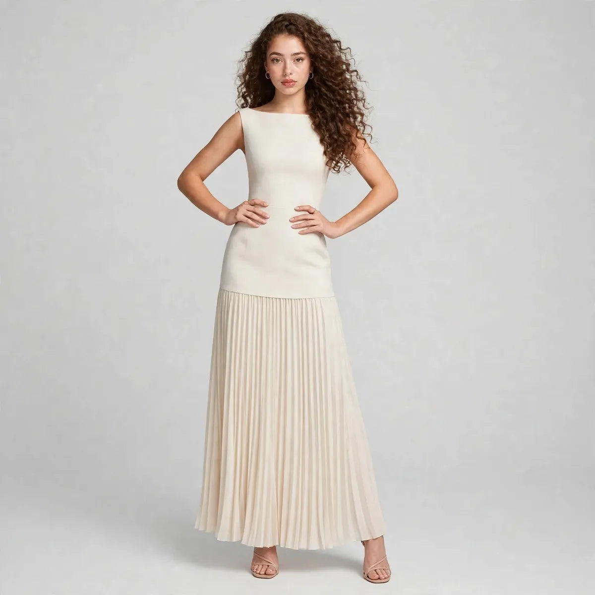 Full-body front view of a cream sleeveless pleated maxi dress with structured bodice, model with hands at waist.