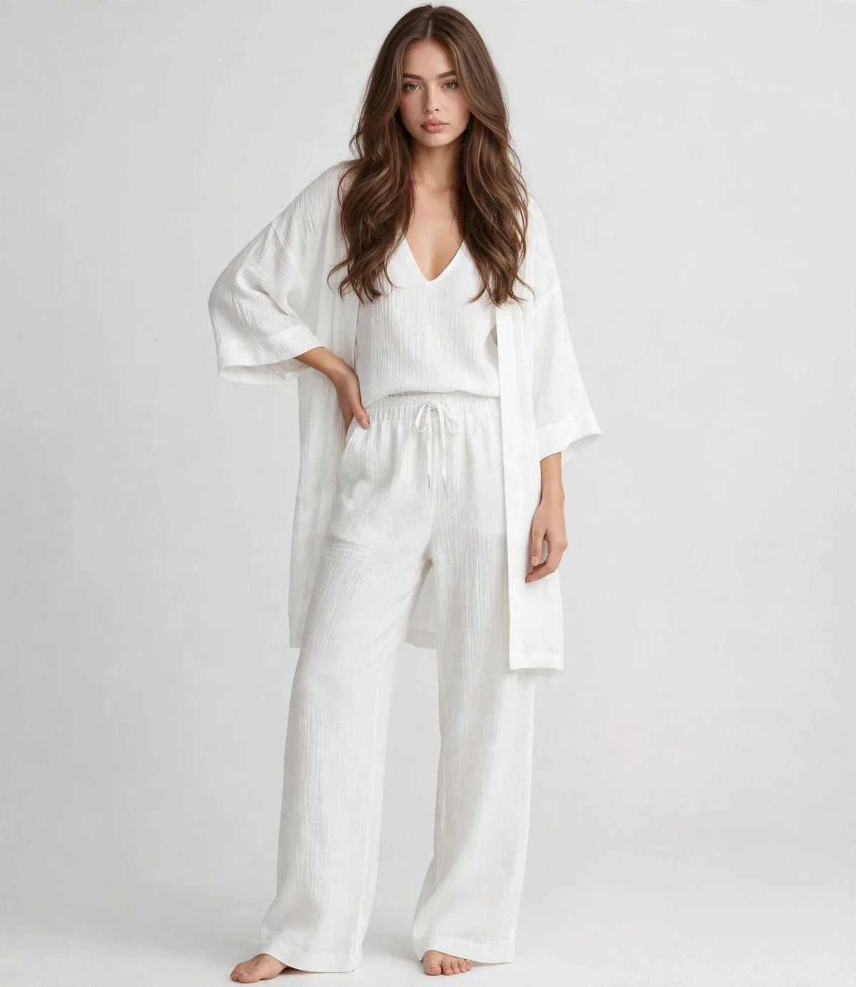 Woman in a white V-neck top, open cardigan and drawstring pants set, standing with hand on hip, gray studio background.
