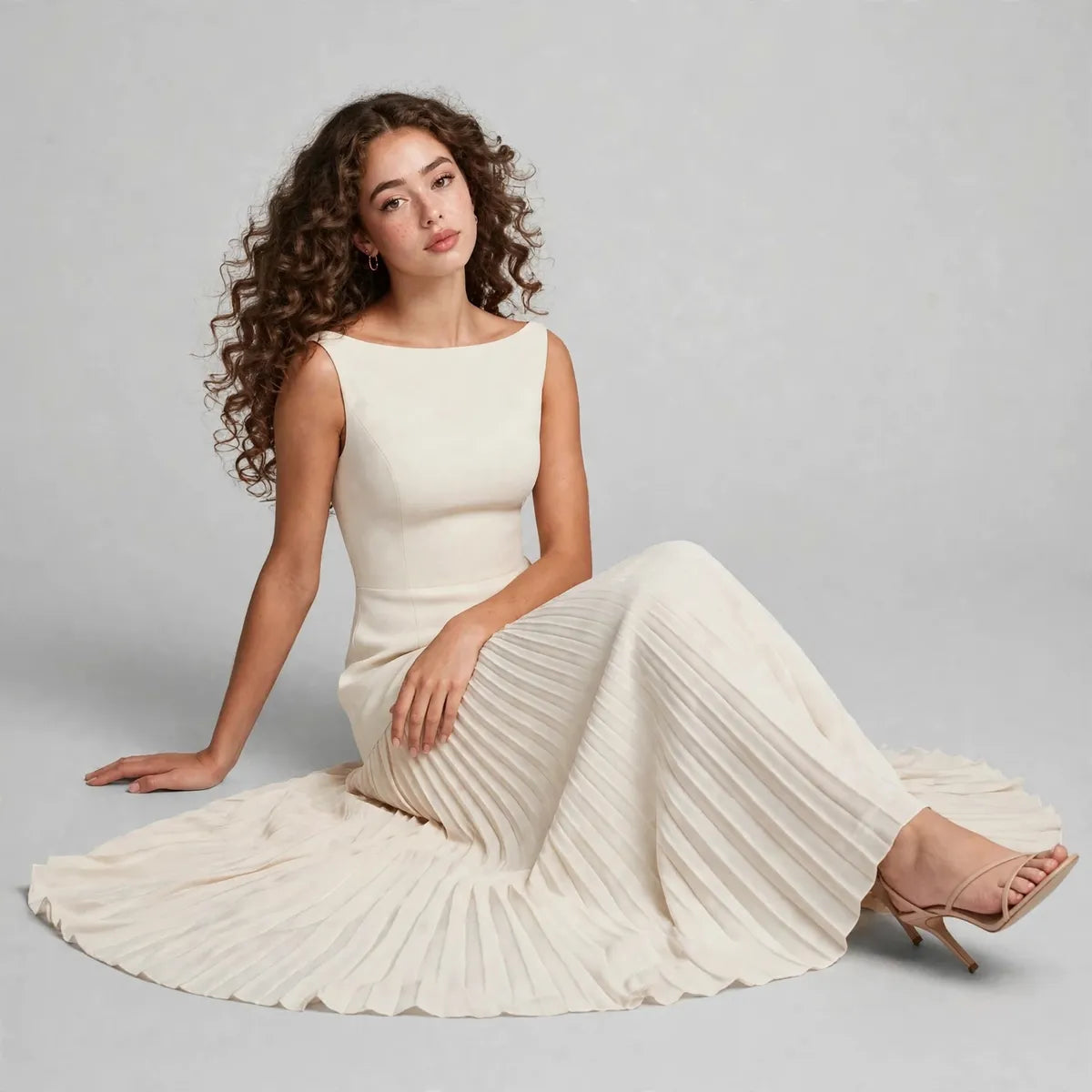 Model seated on a studio floor wearing a cream sleeveless pleated maxi dress with the skirt fanned out around her.