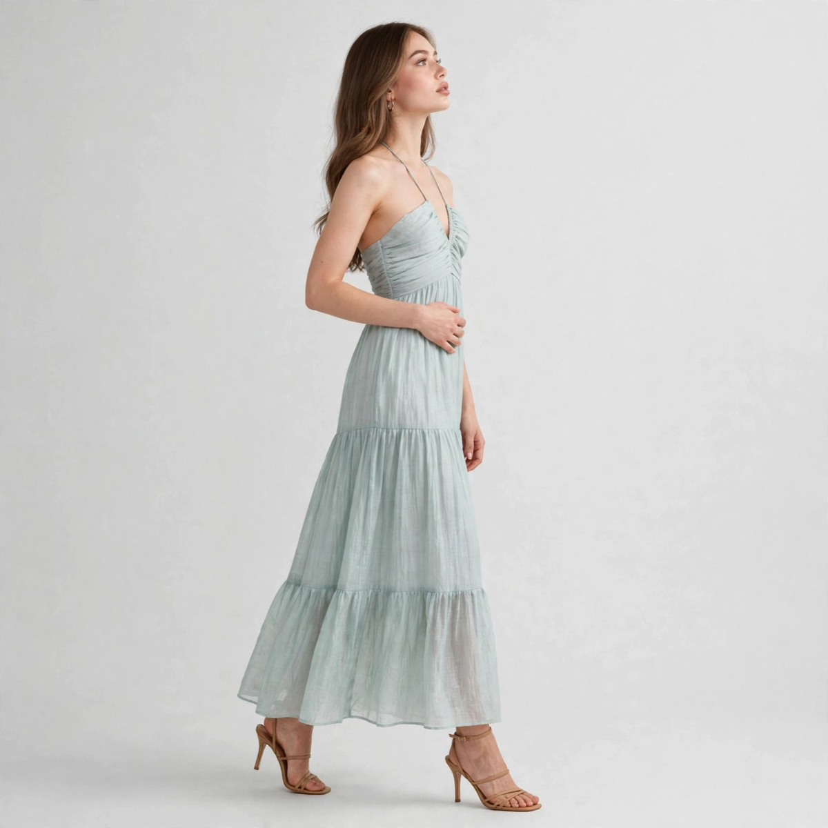 Side view of a woman in a sage green halter-neck tiered maxi dress with tan heels, full-length, gray studio background.