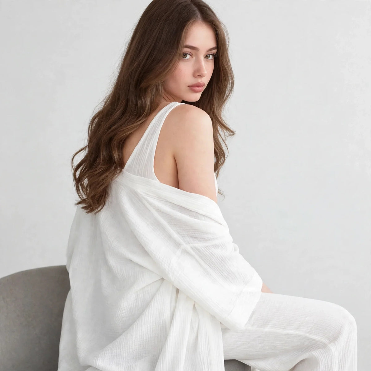 Seated side view of a woman in a white V-neck top and open cardigan set, cardigan slipping off the shoulder, gray background.